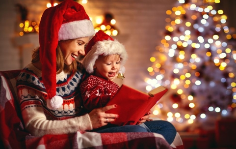A mother and her child sitting by a Christmas tree wearing Santa hats and reading a Christmas book.